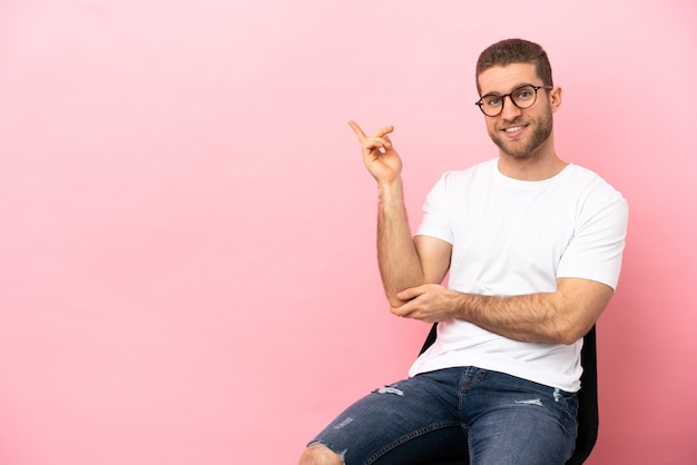 Jovem sentado em uma cadeira sobre um fundo rosa isolado apontando o dedo para o lado