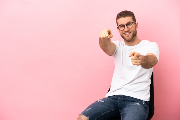 Jovem sentado em uma cadeira sobre um fundo rosa isolado apontando para a frente com uma expressão feliz