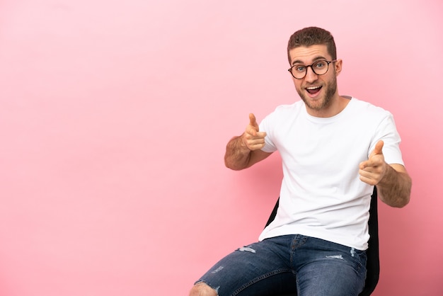 Jovem sentado em uma cadeira sobre um fundo rosa isolado apontando para a frente e sorrindo