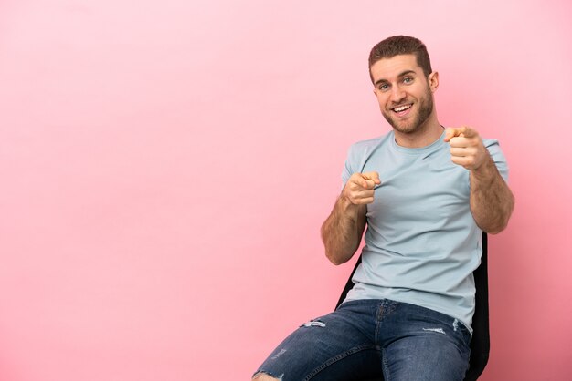 Jovem sentado em uma cadeira sobre um fundo rosa isolado surpreso e apontando para a frente