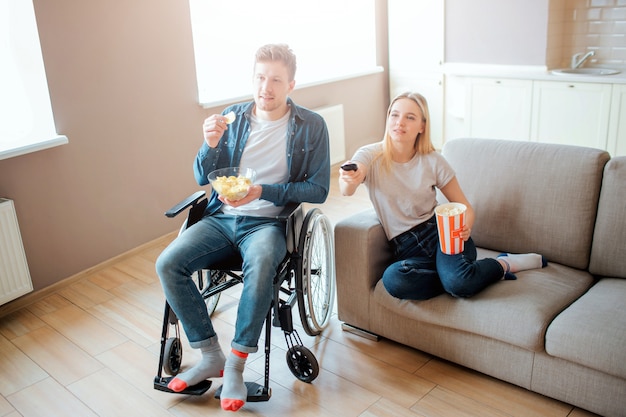 Jovem sentado na cadeira de rodas e assistindo filme com a namorada. cara com deficiência e necessidades especiais. jovem mulher sentar no sofá e segure a tigela com comida. controle remoto.