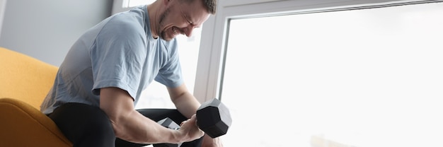 Jovem sentado na cadeira e levantando halteres pesados. Conceito de exercícios esportivos matinais em casa