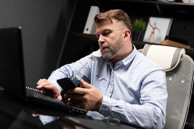 Jovem sentado na mesa do computador e segurando o telefone fixo e celular