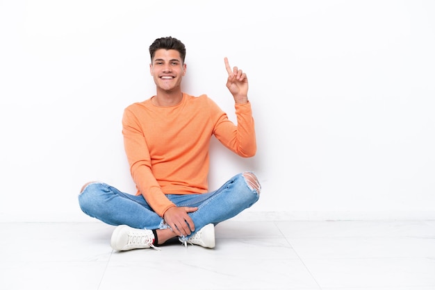 Jovem sentado no chão isolado no fundo branco mostrando e levantando um dedo em sinal do melhor