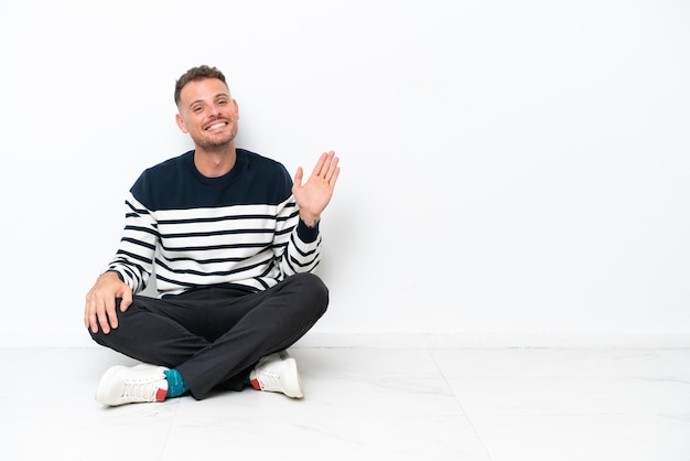 Jovem sentado no chão isolado no fundo branco saudando com a mão com expressão feliz