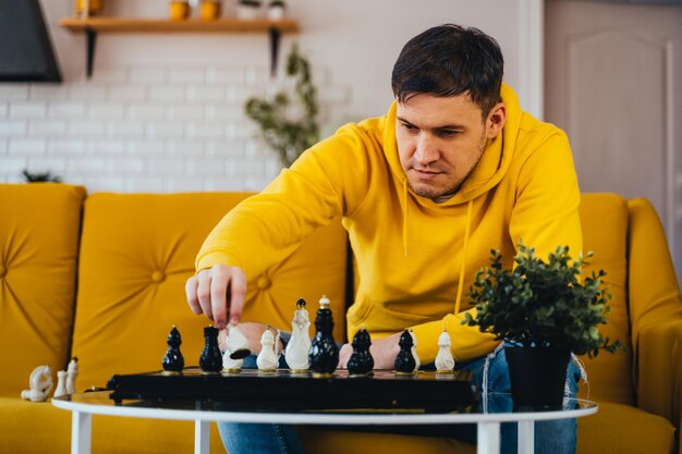 Foto jovem sentado no sofá amarelo e jogando xadrez na sala macho jogando no jogo de tabuleiro lógico consigo mesmo