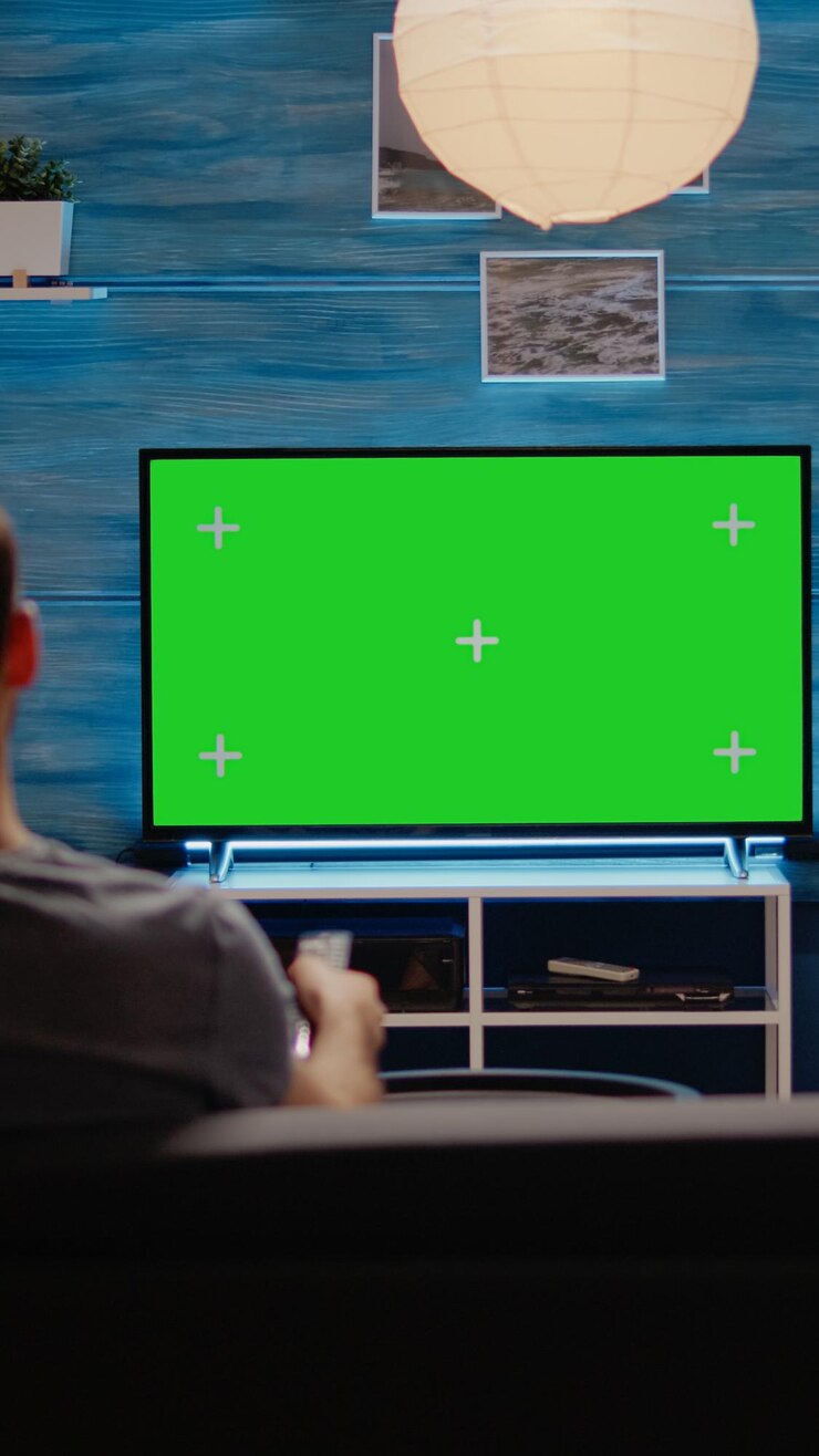 Foto jovem sentado no sofá com tela verde na tv na moderna sala de estar. pessoa usando o controle remoto da televisão com tela de chroma key para modelo isolado de maquete e design de fundo em branco