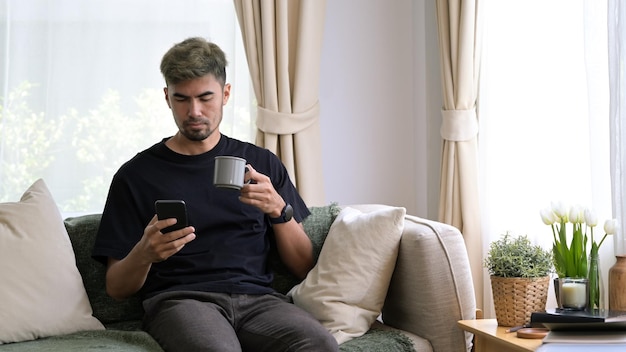 Jovem sentado no sofá confortável e usando telefone inteligente, aproveitando o fim de semana de lazer em casa