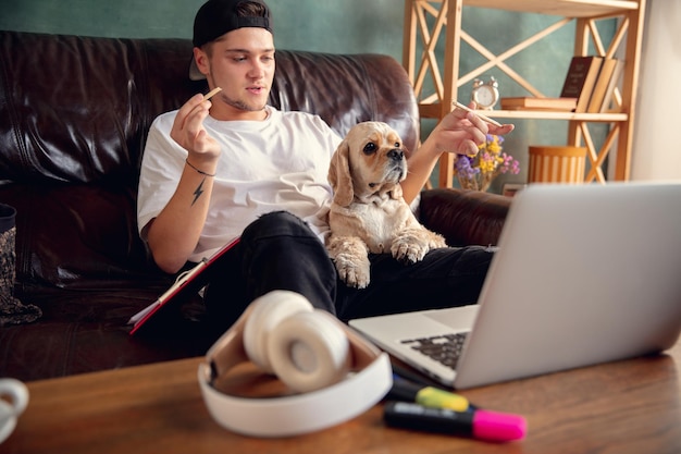 Jovem sentado no sofá em casa com um cachorro fofo e conversando com seus amigos online.