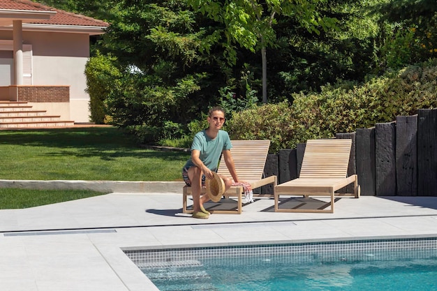 Jovem sentado relaxado em uma espreguiçadeira em sua piscina de jardim em um dia quente de verão