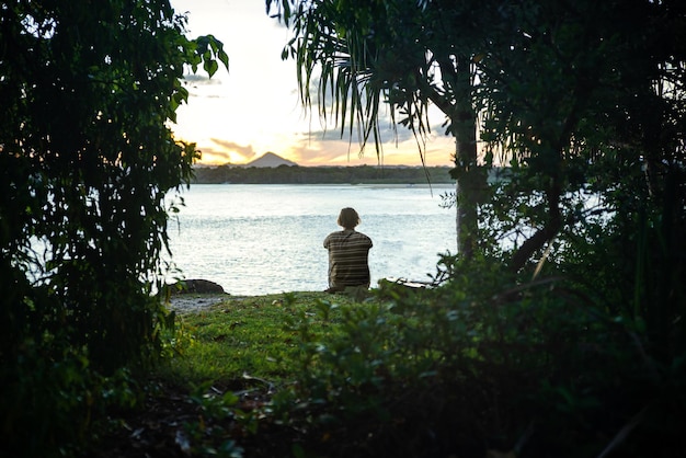 Jovem sentado sozinho no Prado em uma praia olhando o pôr do sol em NoosaQueenslandAustrália