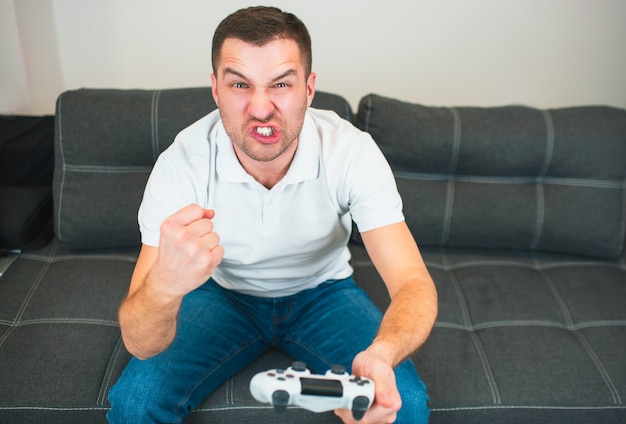 Jovem, sente-se na sala durante a quarentena. Cara jogando intenso jogo de computador sozinho. Mostrando diferentes emoções rudes em seu rosto.