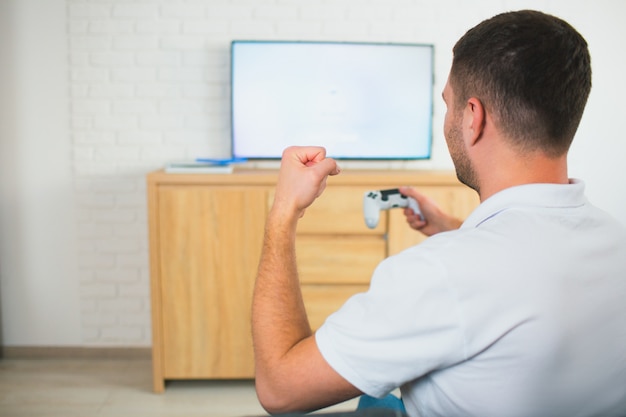 Jovem sente-se na sala jogando tv jogo de computador usando o joystick. Torcendo sozinho no quarto.