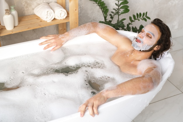 Jovem sereno com máscara facial relaxando em um banho quente com espuma enquanto mantém os olhos fechados