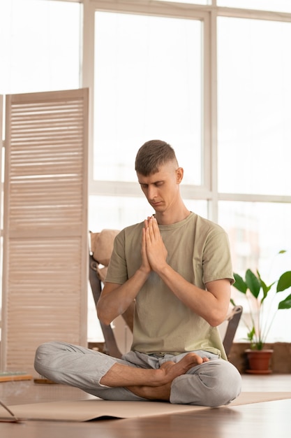 Jovem sereno em roupas esportivas, cruzando as pernas enquanto está sentado na posição de lótus com as mãos unidas pelo peito e praticando exercícios de ioga