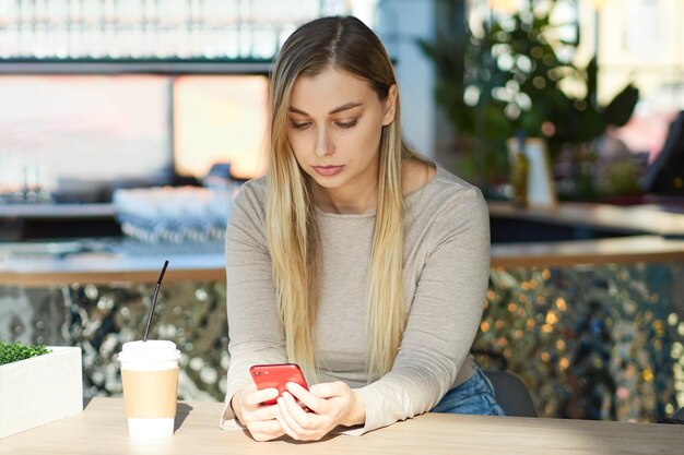 Jovem séria digitando uma mensagem no telefone enquanto está sentado em um café com um copo de papel de bebida