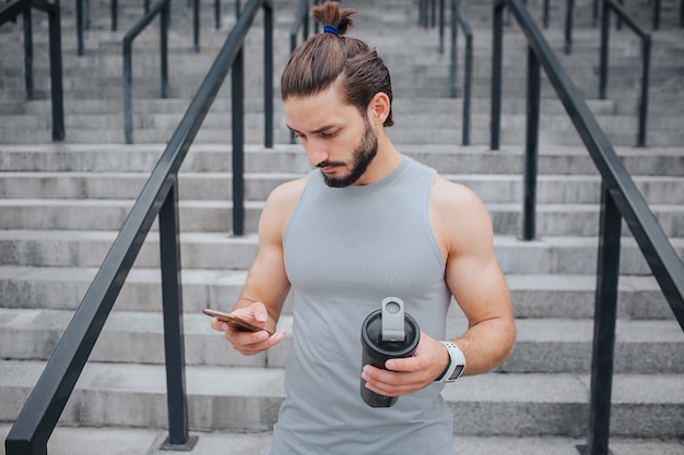 Jovem sério e musculoso fica na escada e olha para o telefone que ele está com uma mão. Há uma garrafa preta em outra. Ele é calmo e concentrado.