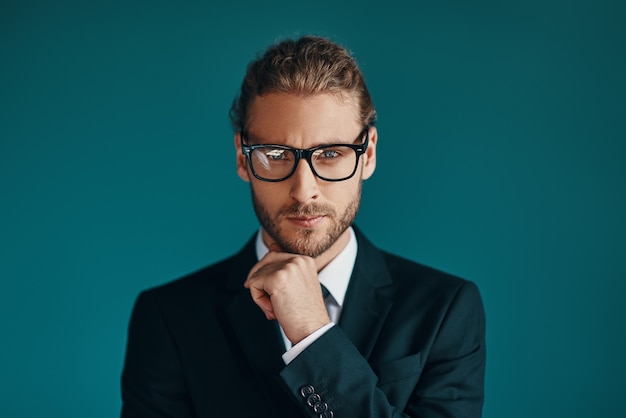 Jovem sério em um terno elegante olhando para a câmera e mantendo a mão no queixo em pé na parede verde