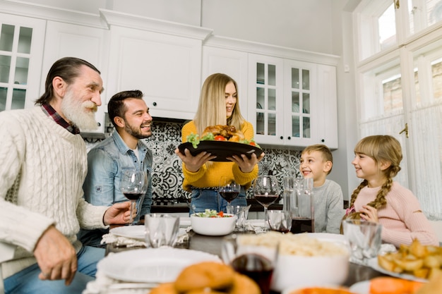 Jovem serve um peru festivo com uma salada, avô, pai e filhos sentar e olhar para a comida saborosa e sorrir