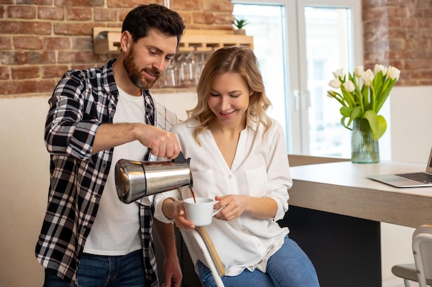 Jovem servindo café à namorada tomando café da manhã na cozinha em casa