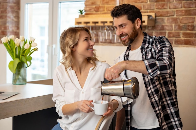 Jovem servindo café à namorada tomando café da manhã na cozinha em casa