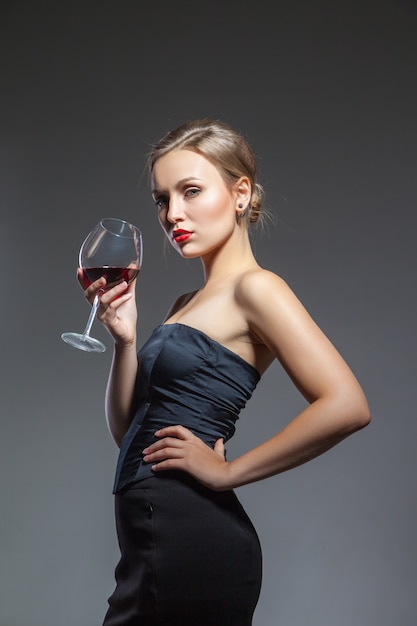 Jovem sexual posando de vestido preto com uma taça de vinho na mão, isolada, sobre o fundo cinza.