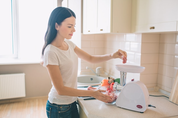 Jovem simpática fica na cozinha. ela mói carne. há uma tigela com vegetais crus além dela.