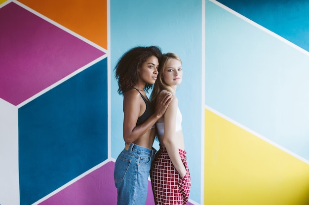 Jovem simpática mulher afro-americana com cabelo escuro e encaracolado e uma linda mulher loira com tops esportivos olhando de lado com uma parede colorida isolada