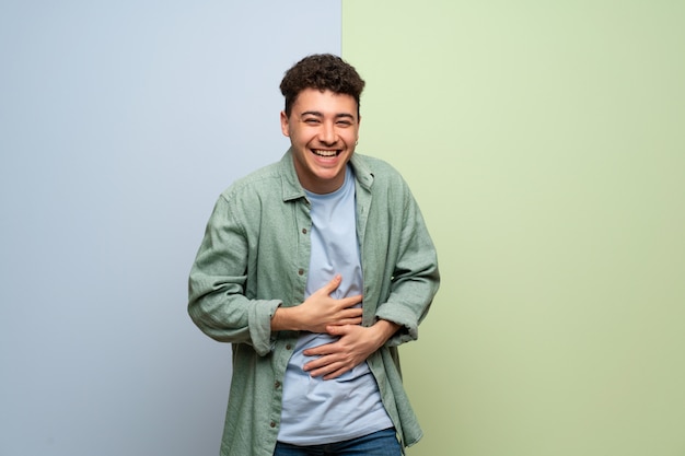 Jovem sobre parede azul e verde, sorrindo muito enquanto coloca as mãos no peito