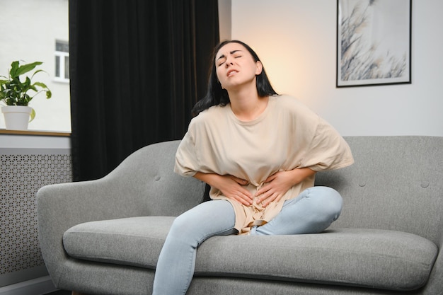 Jovem sofre dor de estômago enquanto está sentada no sofá em casa As causas da dor abdominal incluem dor menstrual gastrite úlcera no estômago intoxicação alimentar diarréia ou IBS Foco seletivo