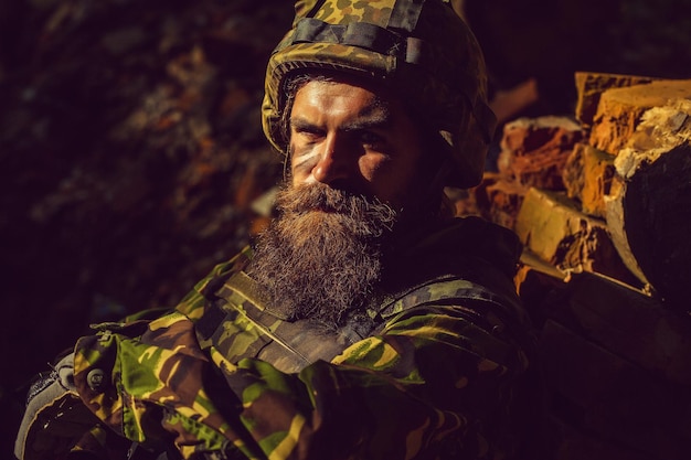 Jovem soldado com a triste cara de barbudo no capacete militar e camuflagem com arma nas costas de tijolos em ruínas ...