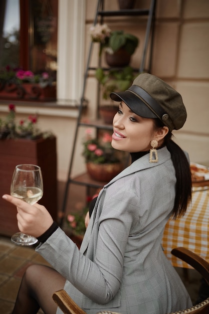 Jovem solteira com roupas da moda de outono sentada sozinha à mesa do café ao ar livre e bebendo vinho