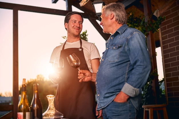 Jovem sommelier alegre com um avental marrom batendo os óculos com um colega mais velho no terraço de uma vinícola