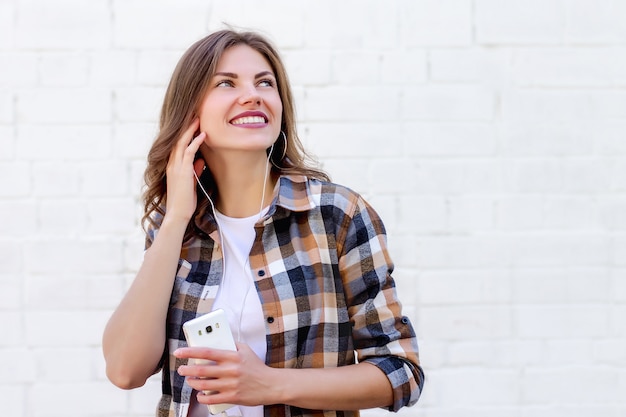 Jovem sorri e ouve música no telefone em um fundo de uma parede de tijolos brancos