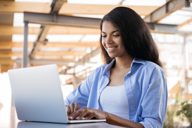 Jovem sorridente afro-americana usando um laptop e trabalhando on-line, sentada em um café