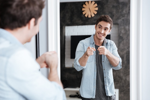 Foto jovem sorridente apontando os dedos para seu reflexo no espelho enquanto está em casa
