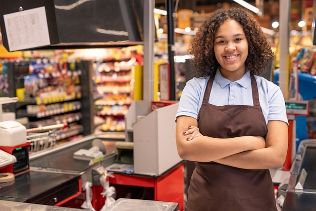 Foto jovem sorridente assistente de loja ou caixa em traje de trabalho, cruzando os braços no peito, enquanto fica de pé no local de trabalho em ambiente de supermercado