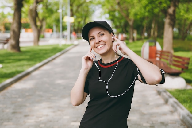 Jovem sorridente atlética linda morena de uniforme preto e boné com fones de ouvido, ouvindo música, mantendo as mãos perto dos ouvidos em treinamento no parque da cidade ao ar livre