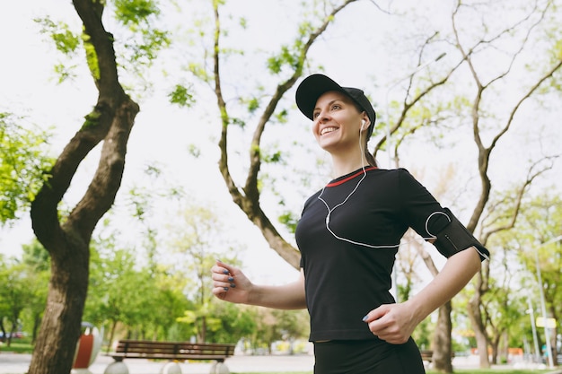 Jovem sorridente atlética mulher morena de uniforme preto e boné com fones de ouvido, treinando, fazendo esporte, correndo, correndo, ouvindo música no caminho no parque da cidade ao ar livre