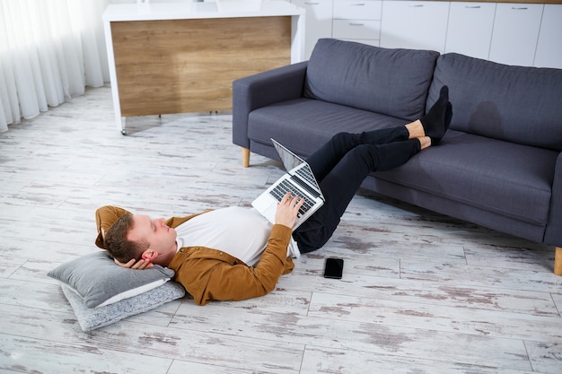 Jovem sorridente atraente e casual sentado no chão em um sofá na sala de estar usando um laptop