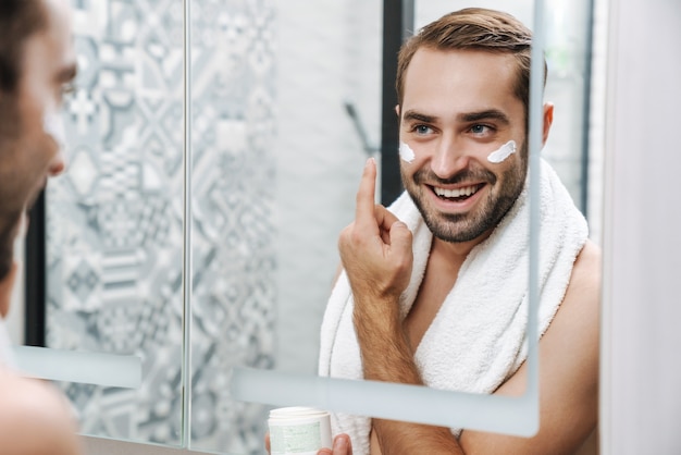 Jovem sorridente atraente sem camisa aplicando creme facial enquanto está em frente ao espelho do banheiro