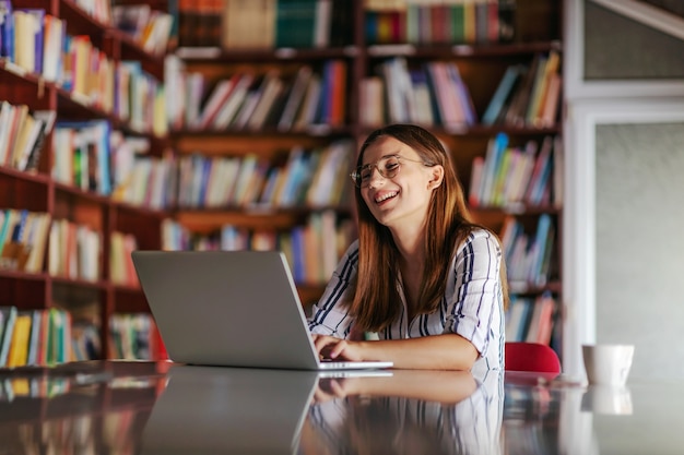 Jovem sorridente atraente universitária sentada na biblioteca e usando o laptop.