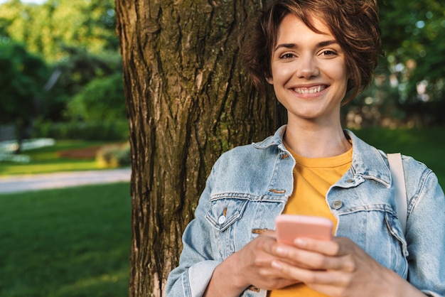 Jovem sorridente atraente, vestindo roupa casual, e passando o tempo ao ar livre no parque, encostado em uma árvore, usando o telefone celular