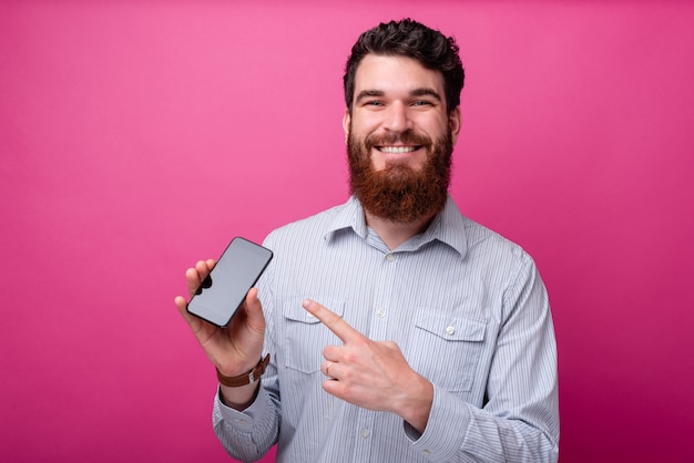 Jovem sorridente barbudo homem mostrando a exibição de um telefone para a câmera no fundo rosa.