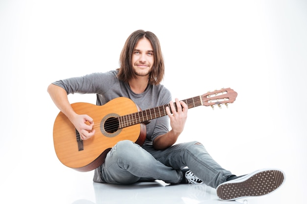 Jovem sorridente com cabelo comprido sentado no chão e tocando violão sobre fundo branco