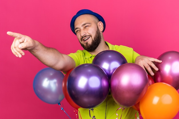 Jovem sorridente com chapéu de festa em pé atrás de balões apontando para o lado isolado na parede rosa