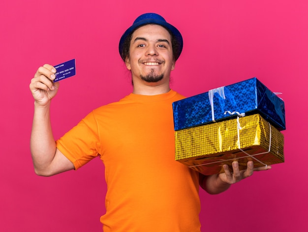 Jovem sorridente com chapéu de festa segurando caixas de presente com cartão de crédito isolado na parede rosa