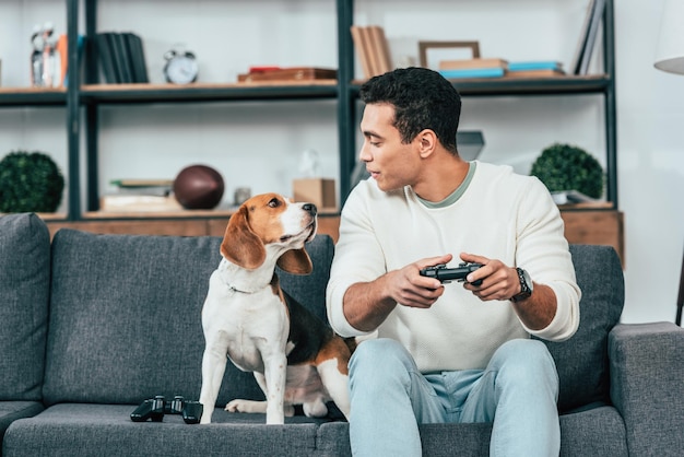 Foto jovem sorridente com gamepad sentado no sofá e olhando para o cachorro