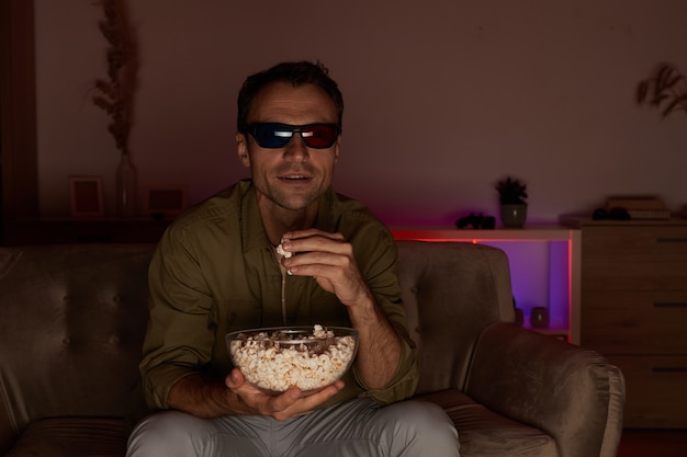 Jovem sorridente com óculos 3d sentado no sofá, comendo pipoca e assistindo filme 3d em casa