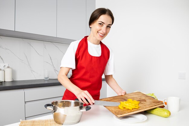 Foto jovem sorridente de avental vermelho cortando legumes na cozinha